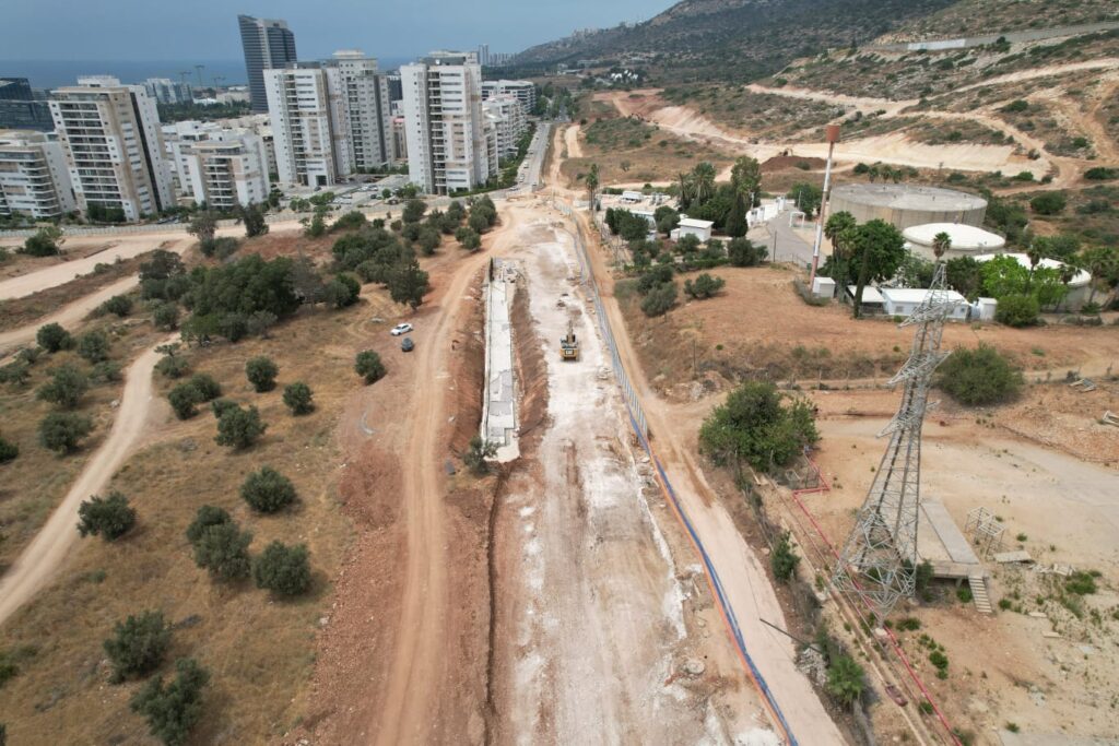 מבט אווירי על אתר בנייה שבו ניכרת ציוד כבד, המוקף בדרכי עפר, עצים, בנייני דירות וגבעות ברקע.