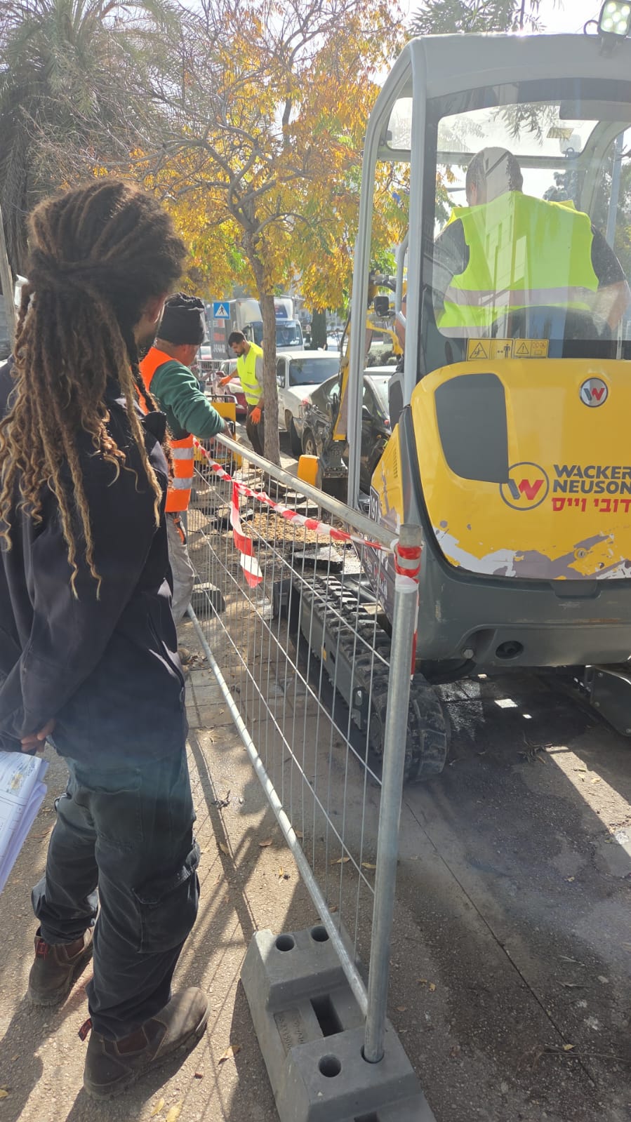 מספר עובדים לבושים באפודי בטיחות פועלים סביב מחפר צהוב קטן על מדרכה עירונית, כשהם מופרדים מהולכי הרגל באמצעות גדר מתכת וסרט אדום.