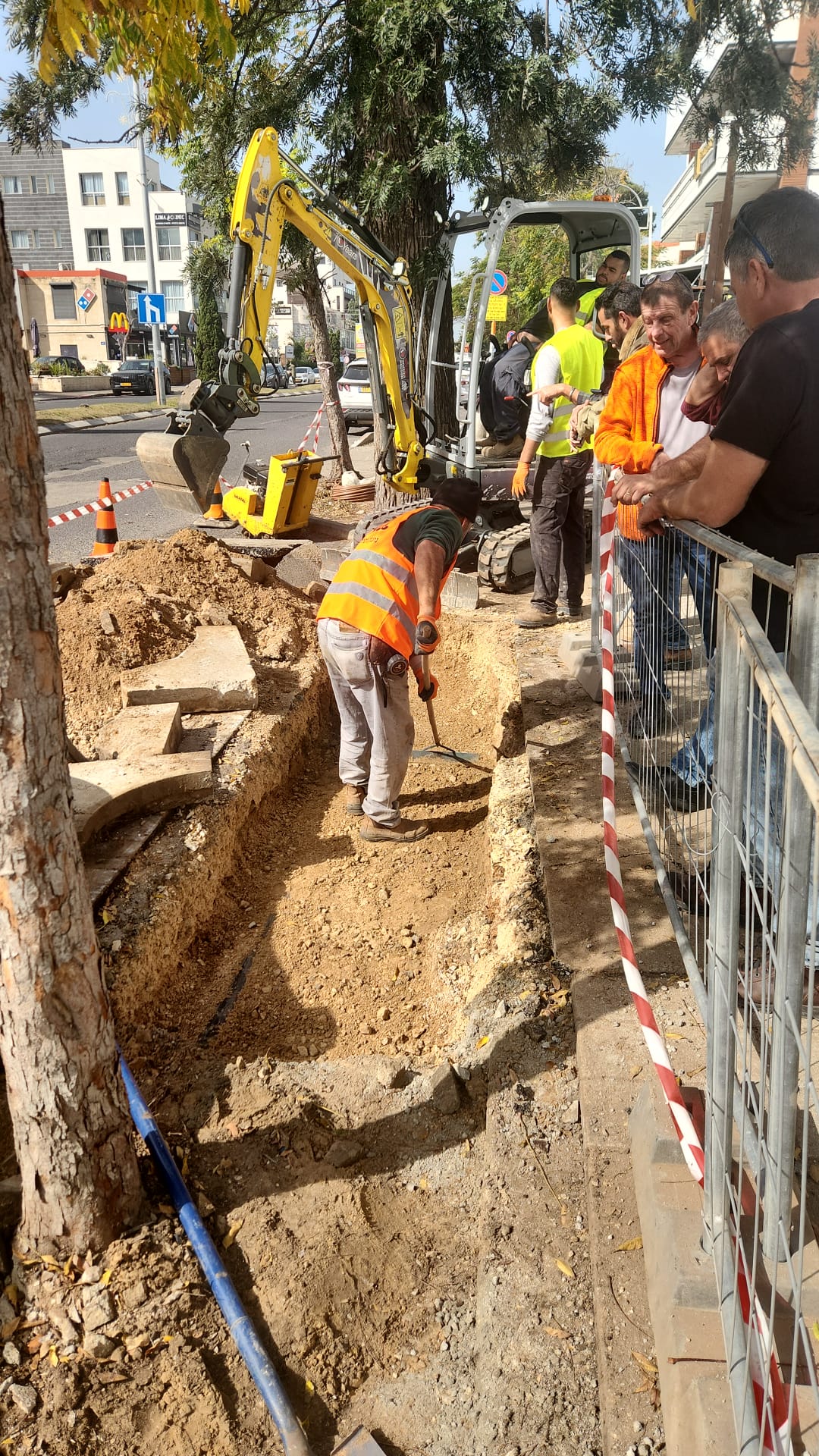 פועלים משתמשים במכונות ובכלי עבודה ידניים כדי לחפור תעלה לאורך מדרכה בעיר. במקום ניכרים מחסומי בטיחות, קונוסים וצופים.