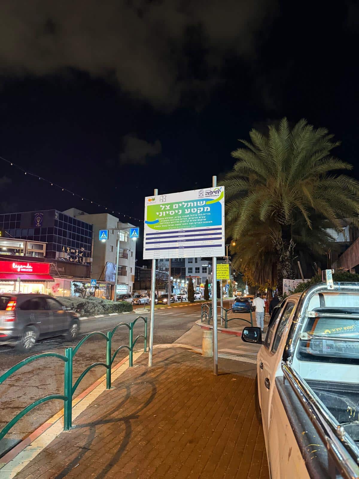 רחוב מואר היטב בלילה, עם מכוניות, חנויות, עץ דקל ושלט גדול בעברית על המדרכה.
