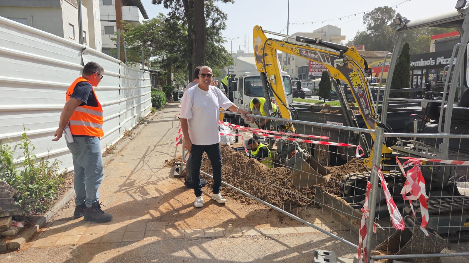 שני גברים עומדים על המדרכה ליד אתר בנייה, כשמאחוריהם, מעבר למחסום בטיחות ולסרט אדום-לבן, נראים פועלים ומחפר.