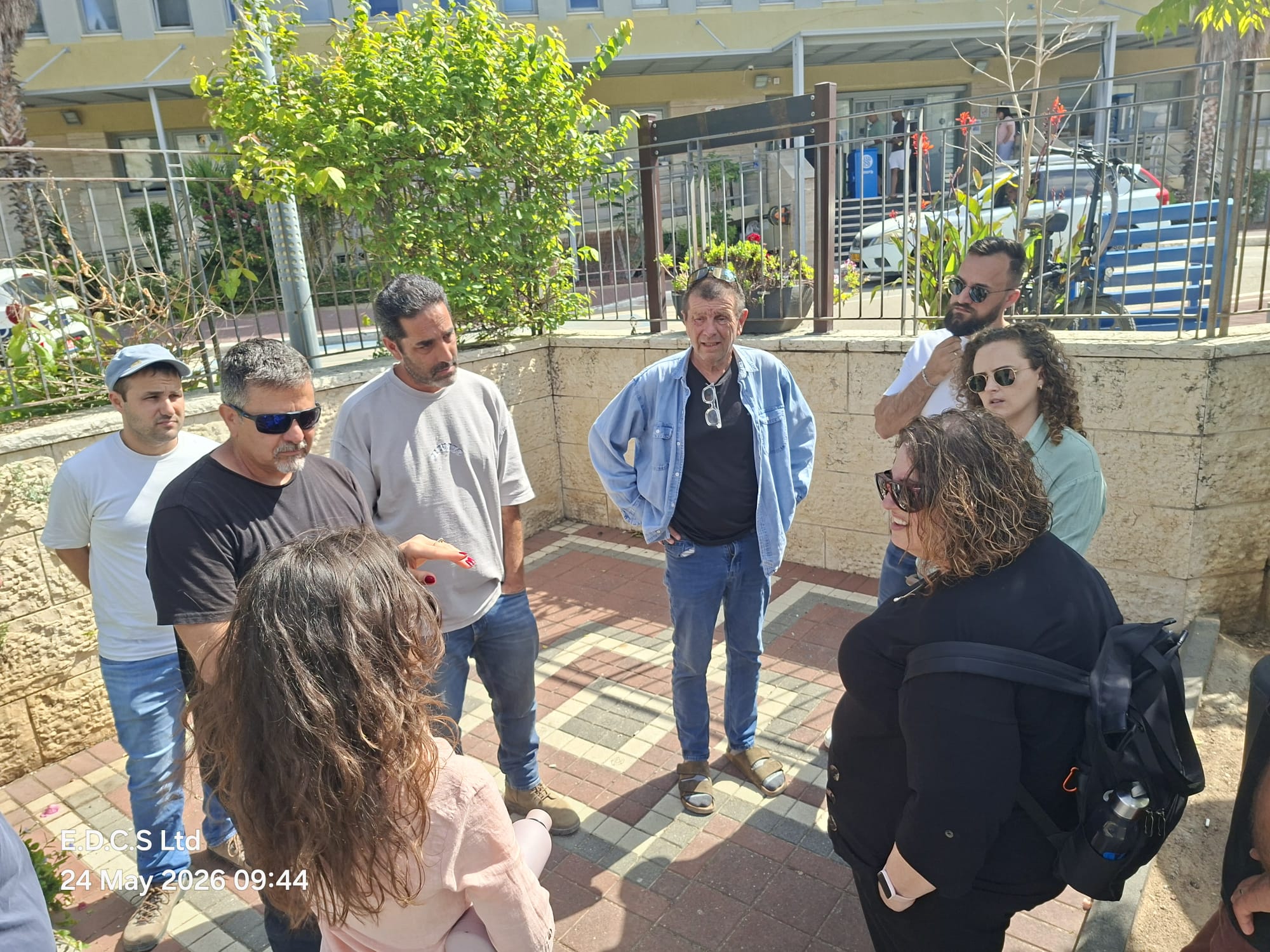 קבוצת אנשים עומדת בחוץ במעגל חצי-עגול, משוחחת ליד קיר אבן וגדר מתכת, תחת שמש זורחת.