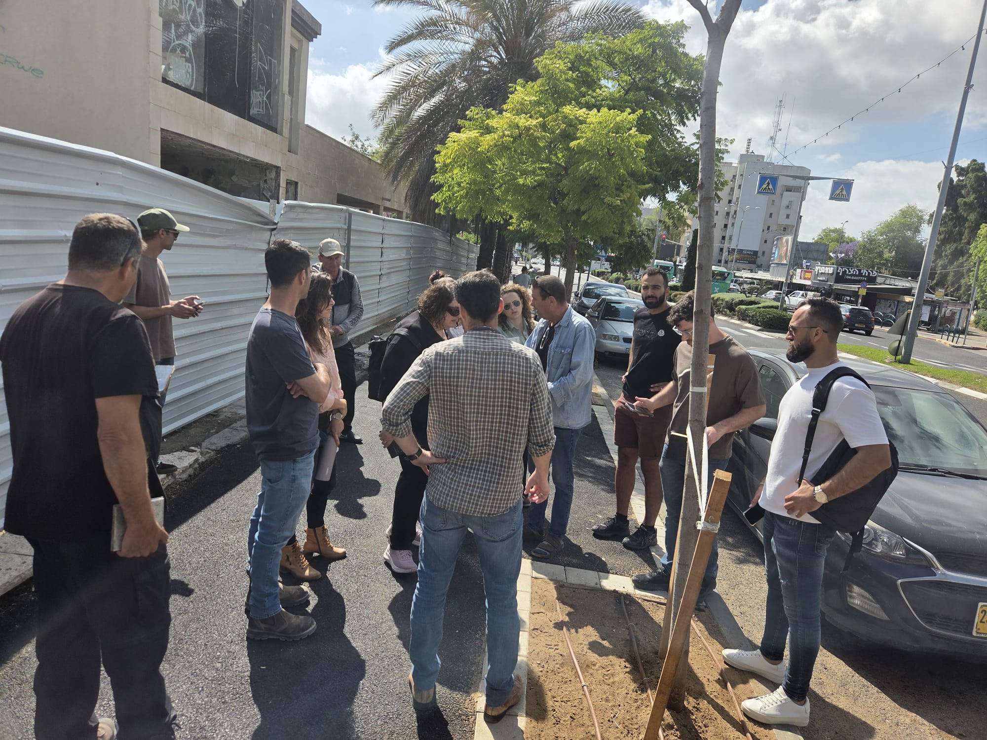 קבוצת אנשים עומדת יחד על מדרכה שטופת שמש ליד אתר בנייה; חלקם משוחחים ואחרים מקשיבים, כשברקע נראים מכוניות ועצים.