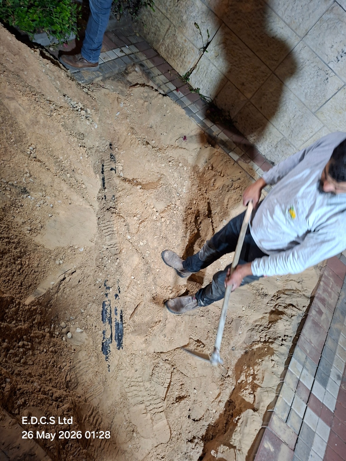 אדם המשתמש בכף כדי לחפור באדמה חולית ליד שביל מרוצף בשעת לילה. בחלק העליון נראית דמותו של אדם נוסף. התמונה צולמה ב-26 במאי 2026, בשעה 01:28.