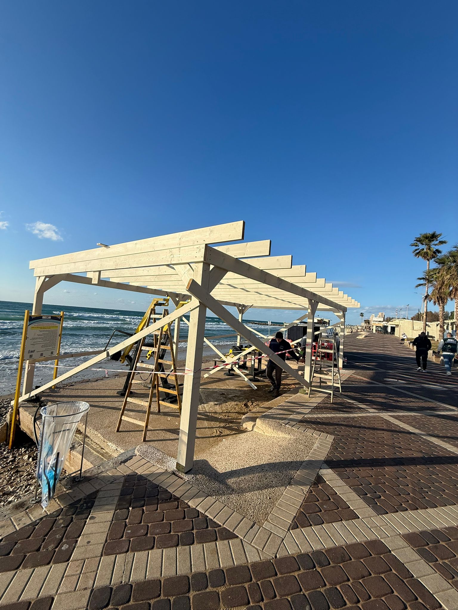 פועלים מרכיבים מבנה עץ על שפת הים, על טיילת מרוצפת בקרבת האוקיינוס, כשניתן להבחין בסולמות ובכלי בנייה.