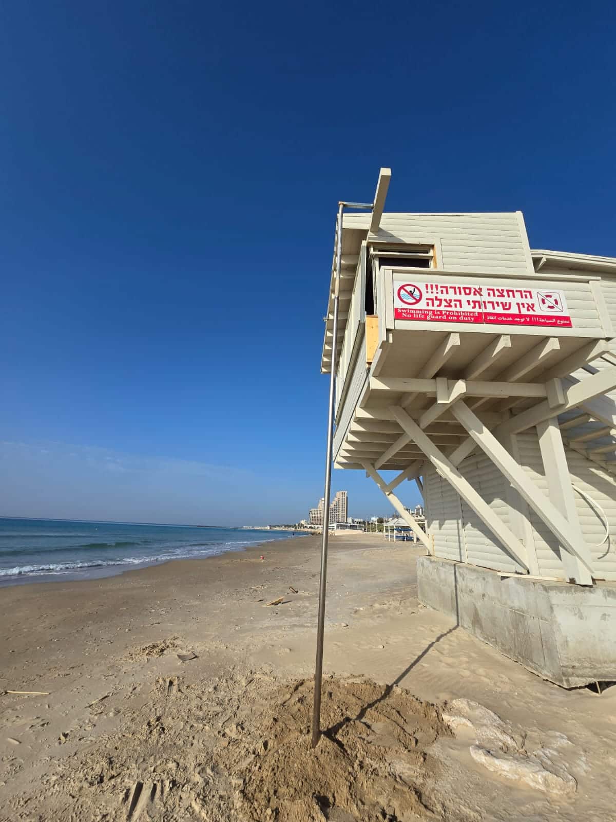 תחנת מצילים בחוף חולי עם שלט בעברית; ניתן לראות את הים ואת הבניינים המרוחקים תחת שמיים כחולים וצלולים.