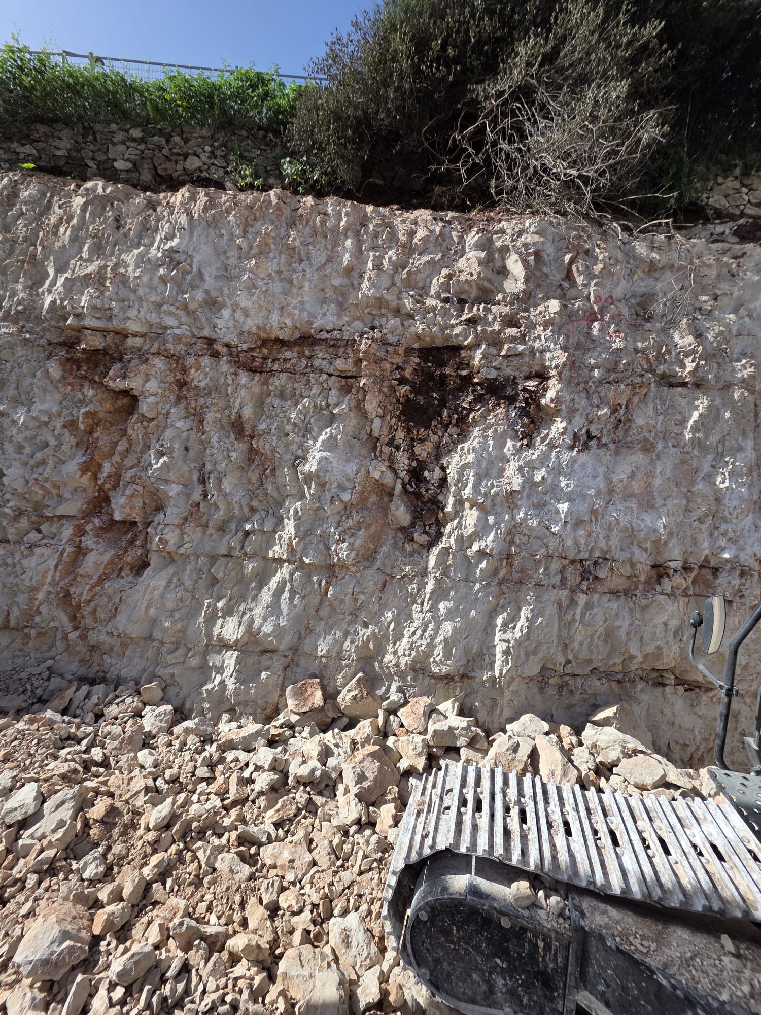 מצוק סלעי עם אבנים רופפות בבסיסו; בפינה הימנית התחתונה ניתן להבחין בחלק ממסלול מתכת של רכב בנייה. שיחים צומחים בראש הקיר הסלעי.