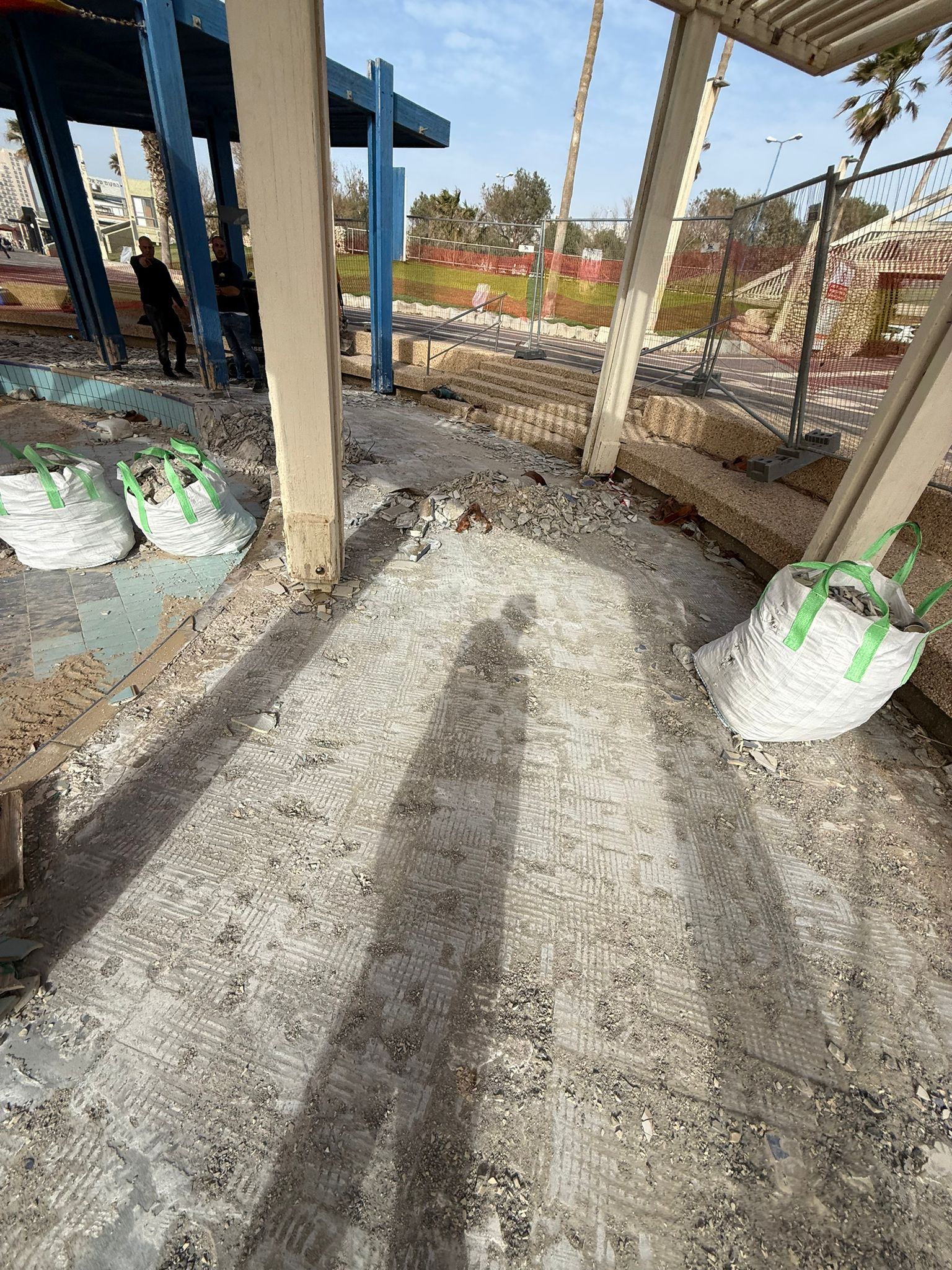 צל של אדם באתר בנייה מאובק, כשברקע שקים גדולים של חומרי בניין, עמודים, גדרות ועצים.