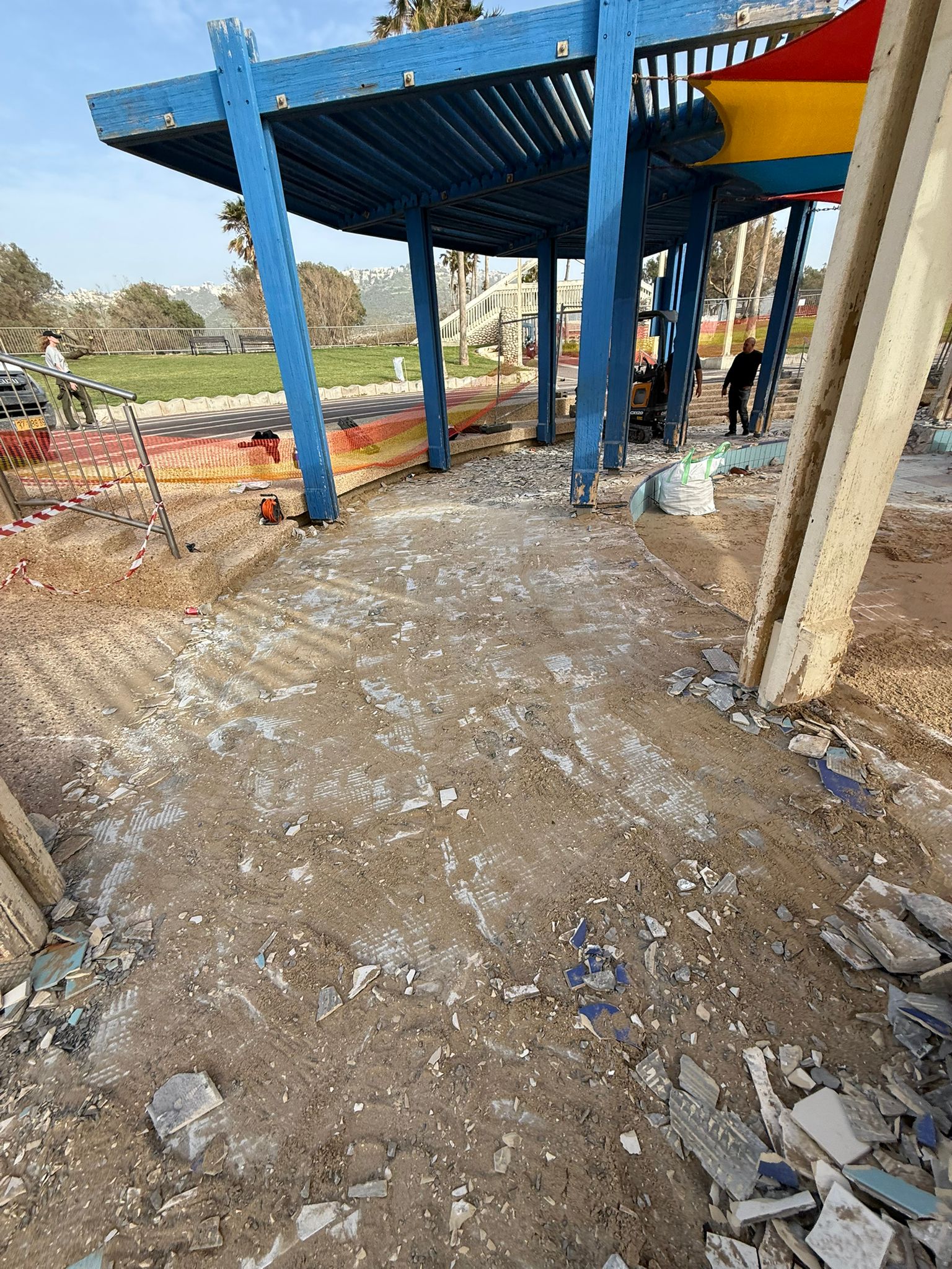 אזור משחקים תחת סככת צל כחולה, עם אריחים שבורים ופסולת על הקרקע; נראים חלקים מהגדר וחומרי בנייה; ברקע נראים שני אנשים עובדים.