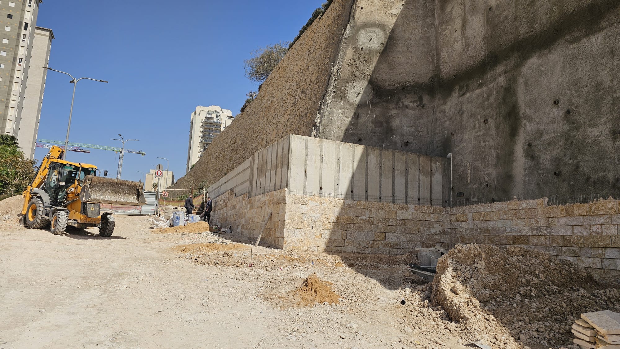 אתר בנייה שבו נראית מחפרת-מעמיסה, פועלים, ערימות עפר וקיר תומך מאבן שבנייתו טרם הושלמה, בסמוך לבניינים גבוהים תחת שמיים כחולים וצלולים.