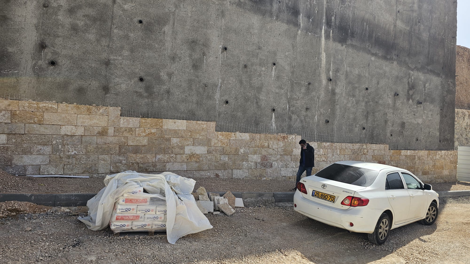 גבר עובר ליד קיר אבן שבנייתו טרם הושלמה, ליד מכונית לבנה חונה וחומרי בנייה מכוסים על משטח עפר.