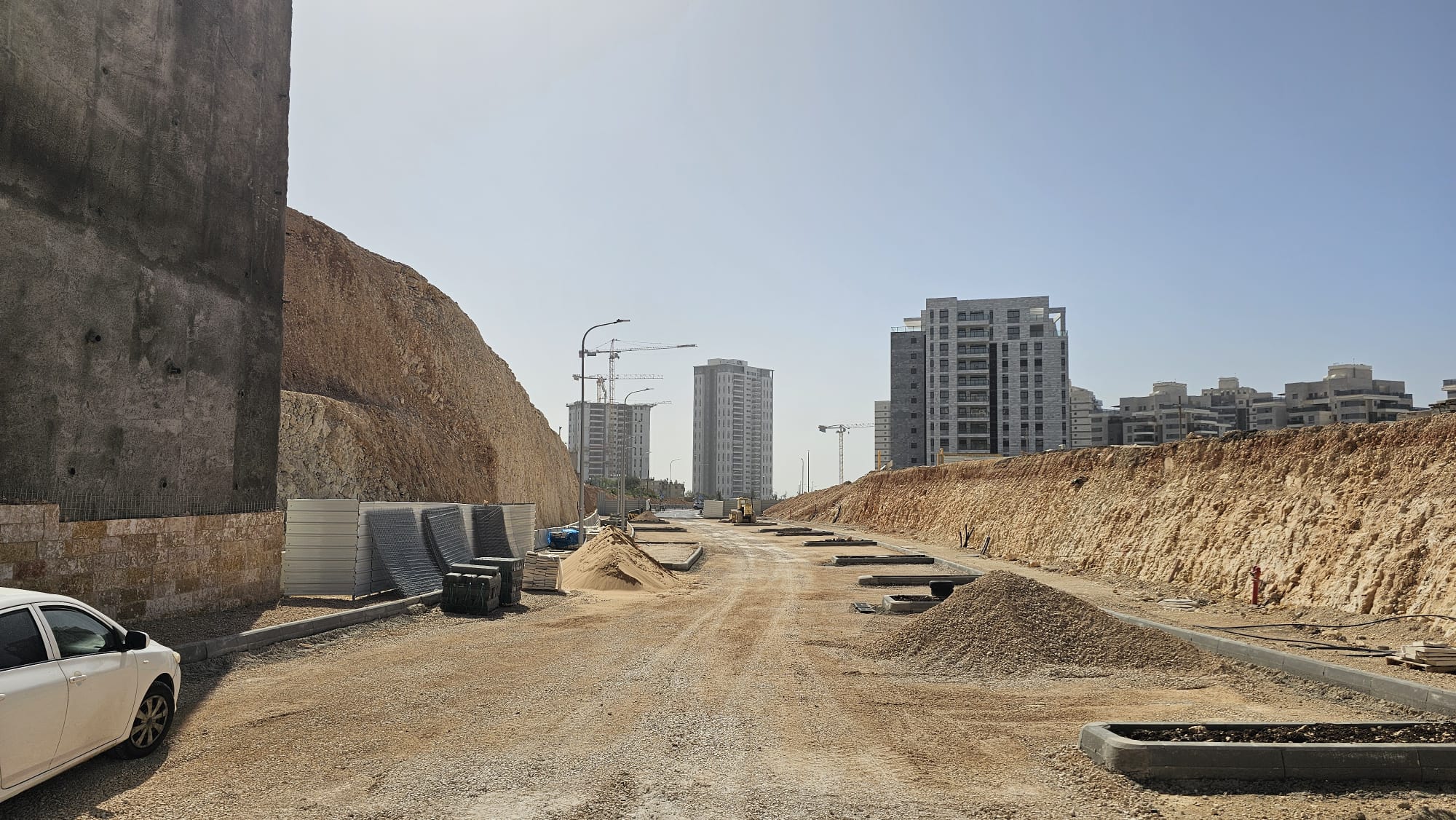 דרך עפר רחבה הנמצאת בבנייה, משני צדיה תלוליות עפר וחומרי בנייה, וברקע בנייני דירות מודרניים תחת שמיים בהירים.