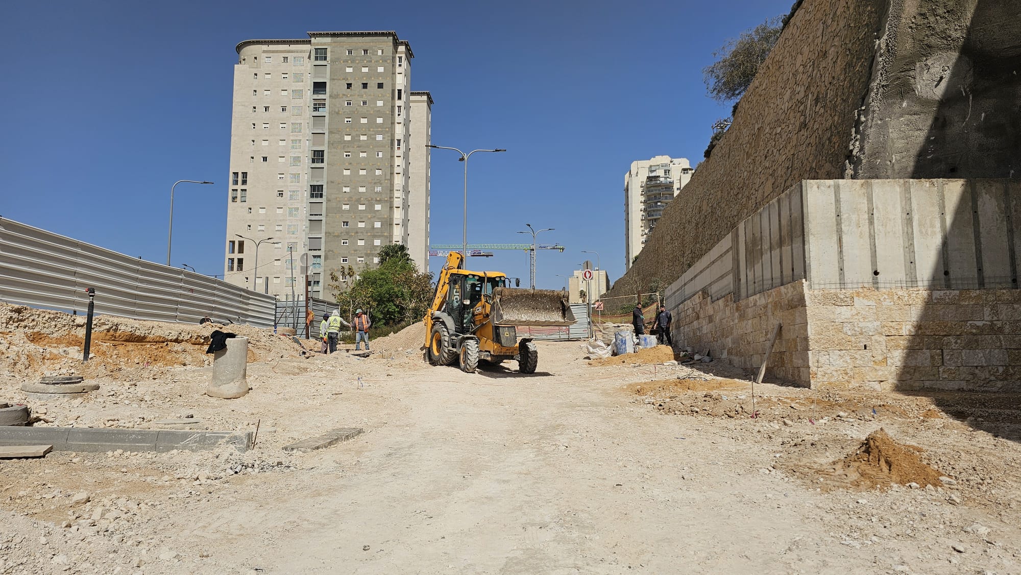 אתר בנייה שבמרכזו עומדת מחפרת-מעמיסה צהובה, מוקפת בפועלים, עפר ובניינים גבוהים ברקע.