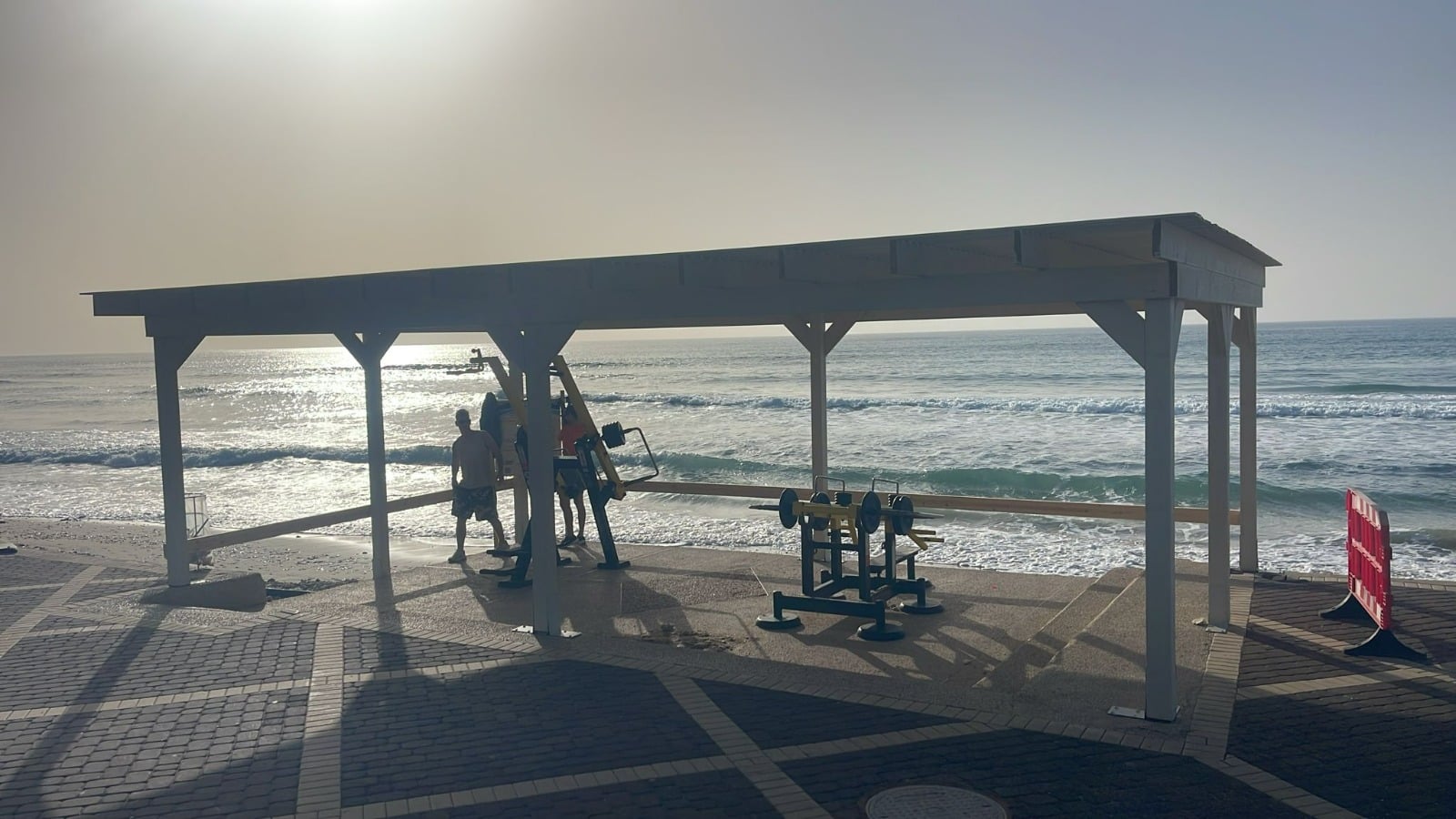 מתחם כושר חיצוני מוצל משקיף על האוקיינוס. כמה אנשים מתאמנים במכשירי הכושר, בעוד קרני השמש מנצנצות על המים ועל הרצפה המרוצפת.