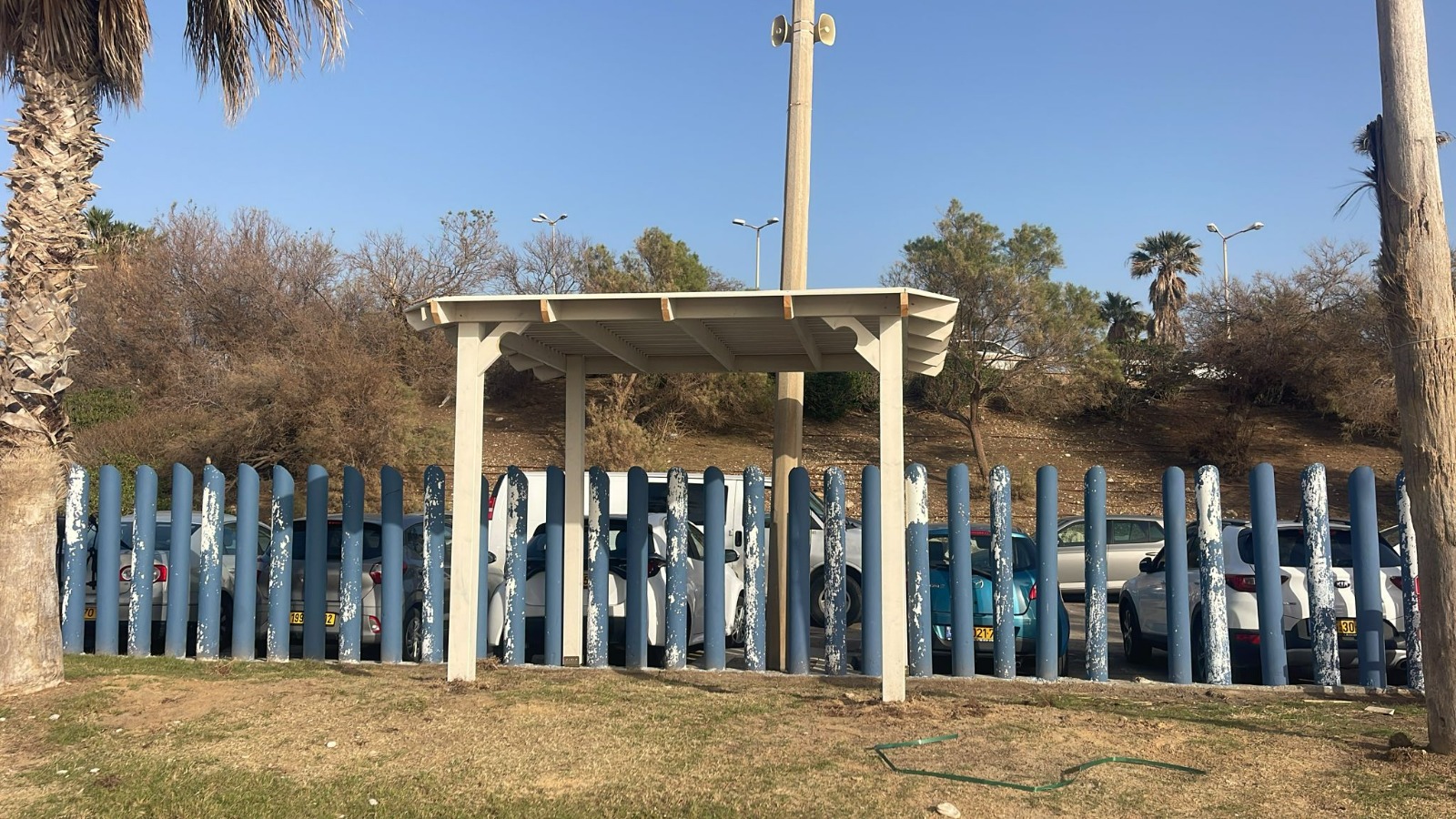 מחסן עץ קטן וגדר כחולה ניצבים מול מכוניות חונות, כשברקע עצי יבש ושמיים בהירים.