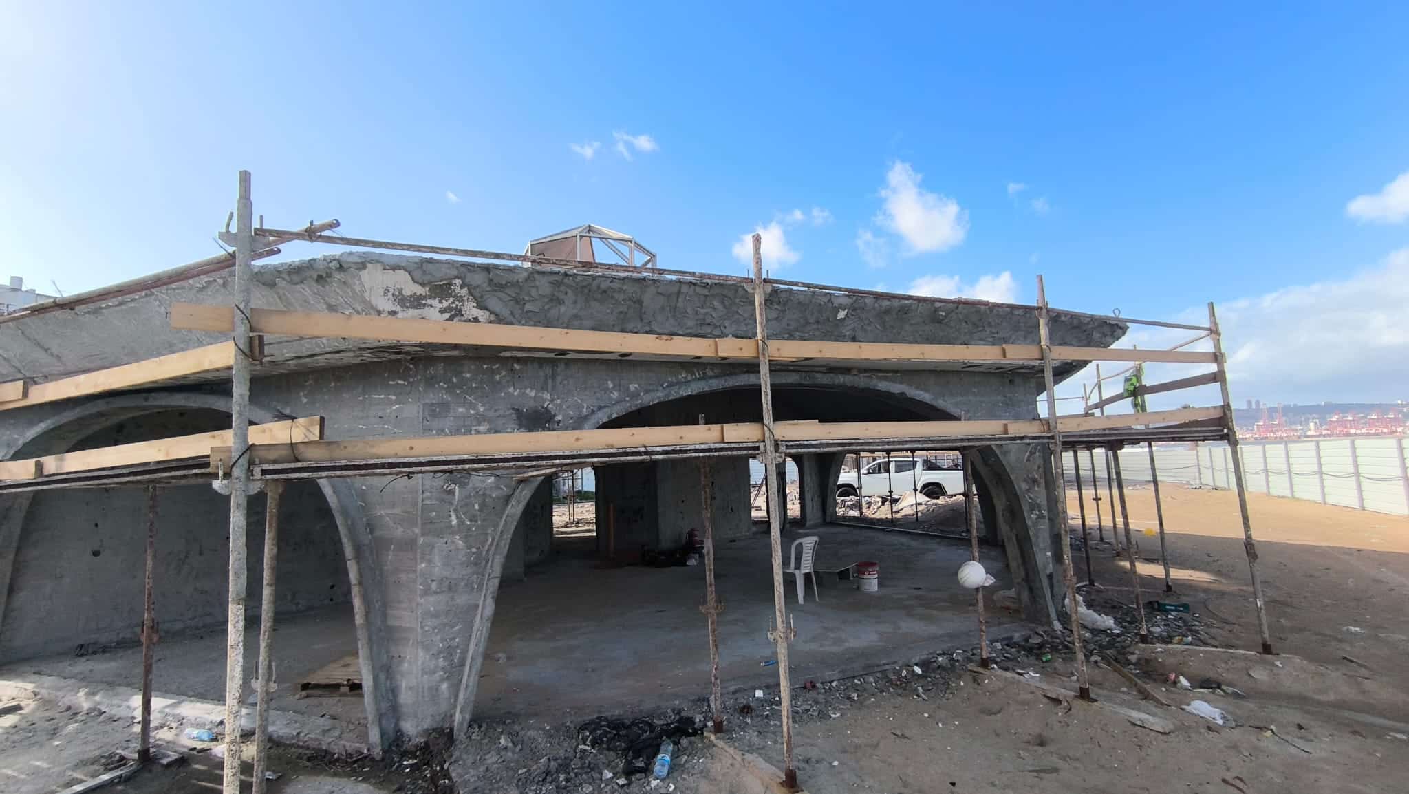 בניין בטון הנמצא בבנייה, עם פיגומים ופסולת, הכולל תומכים מקושתים וגג לא גמור, על רקע שמיים כחולים עם עננים פזורים.