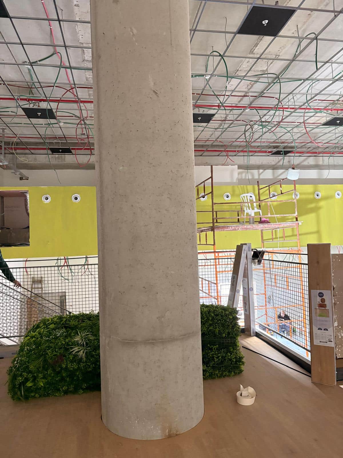 עמוד בטון גדול ניצב בתוך החדר לצד צמחייה וסולם, כשברקע נראים חוטי חשמל התלויים מהתקרה ופיגומים.