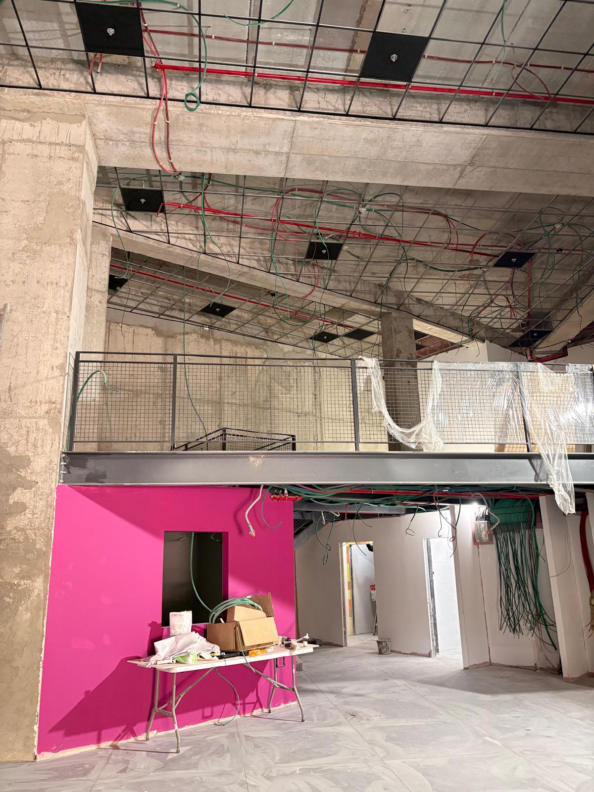 אתר בנייה פנים עם תקרת בטון חשוף, חיווט גלוי, קיר ורוד בוהק, קומת ביניים עם מעקה, ושולחן עם חומרי בנייה בקדמת התמונה.