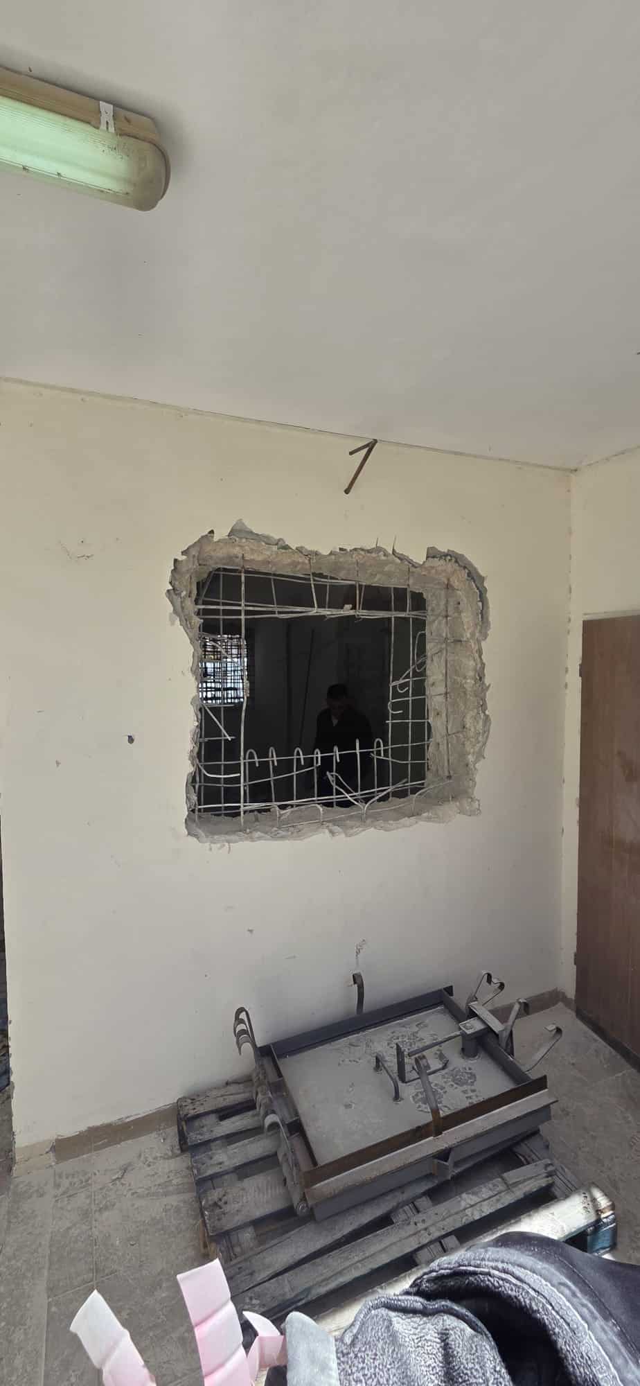 בקיר נפרץ חור מלבני גדול שבו נראים מוטות מברזל חשופים; על הרצפה ליד הפתח מונחים שברי חפצים ושולחן הפוך.