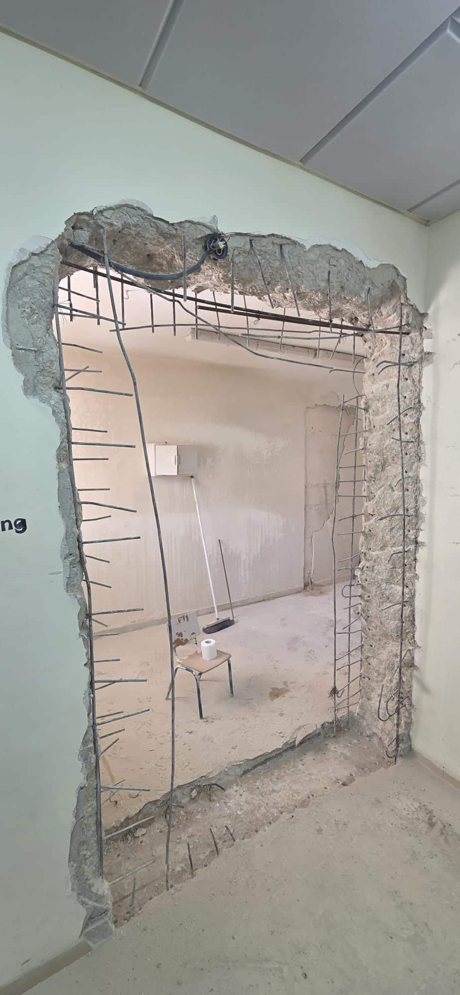 בקיר הבטון נחתך חור גס ומלבני, שבו נראים מוטות מברזל חשופים, וחושף חדר ריק שבתוכו נראים כלי בנייה.