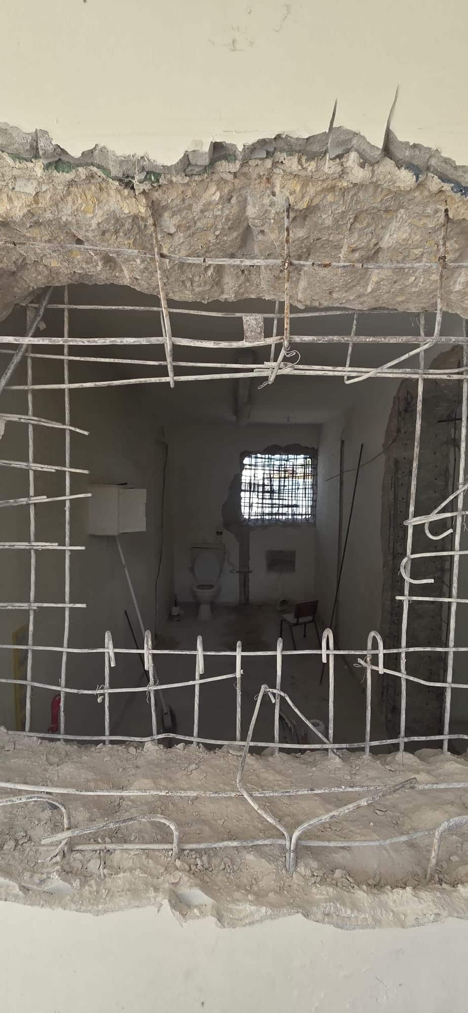 חור גדול בקיר בטון, שבו נראים מוטות מברזל חשופים, חושף חדר ובו שירותים, חלון מסורג ופסולת בנייה.