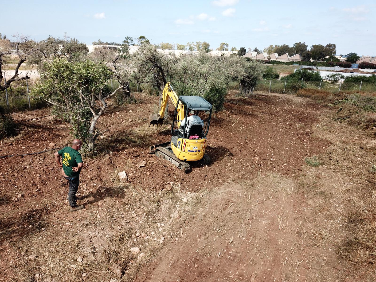 מחפר קטן חופר בשטח עפר שנפנה, בעוד אדם עומד בסמוך. סביב אתר העבודה נראים עצים ושיחים, וברקע נראים בתים.