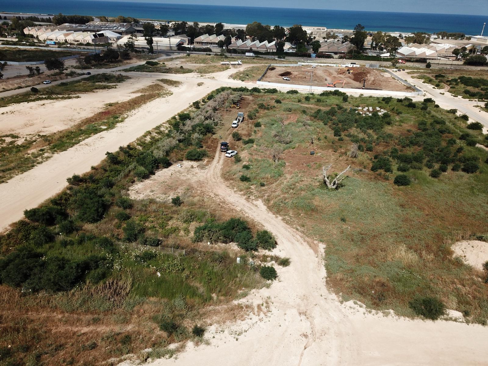 מבט אווירי על אתר בנייה חולני עם צמחייה פזורה, מכוניות חונות ומבנים ברקע, בקרבת קו החוף.