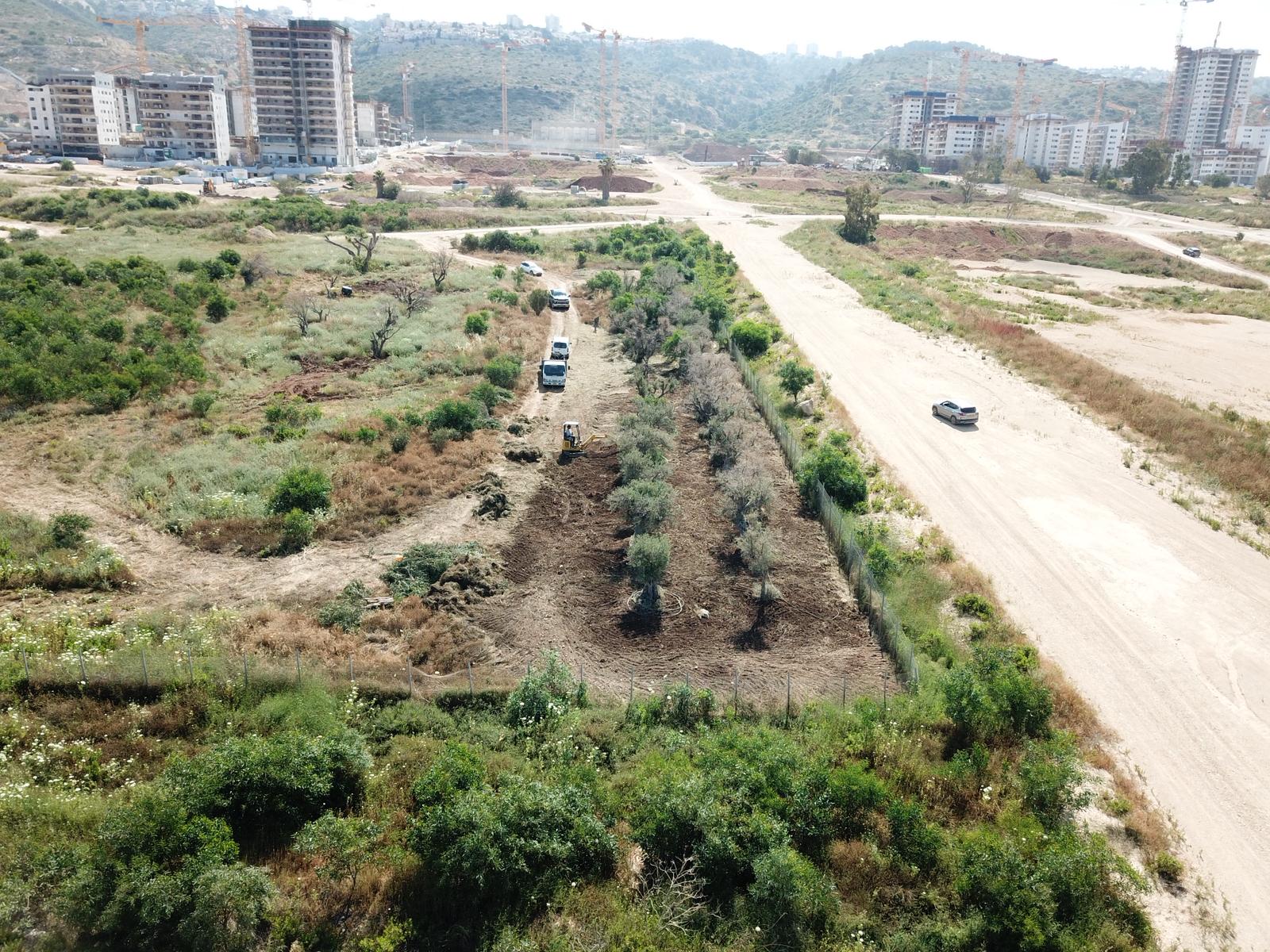 מבט אווירי על אתר בנייה שבו נראים מספר כלי רכב ליד דרך עפר, מוקף בצמחייה, וברקע בניינים רבי קומות הנמצאים בשלבי בנייה חלקיים.