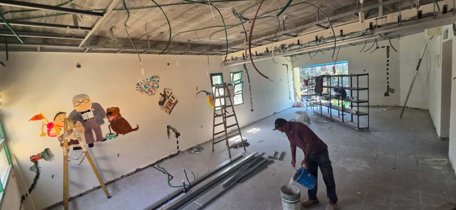 אדם עובד עם חומרי בניין בחדר שבנייתו טרם הושלמה, שבו נראים חוטי תקרה חשופים, יצירות אמנות על הקירות וסולמות.