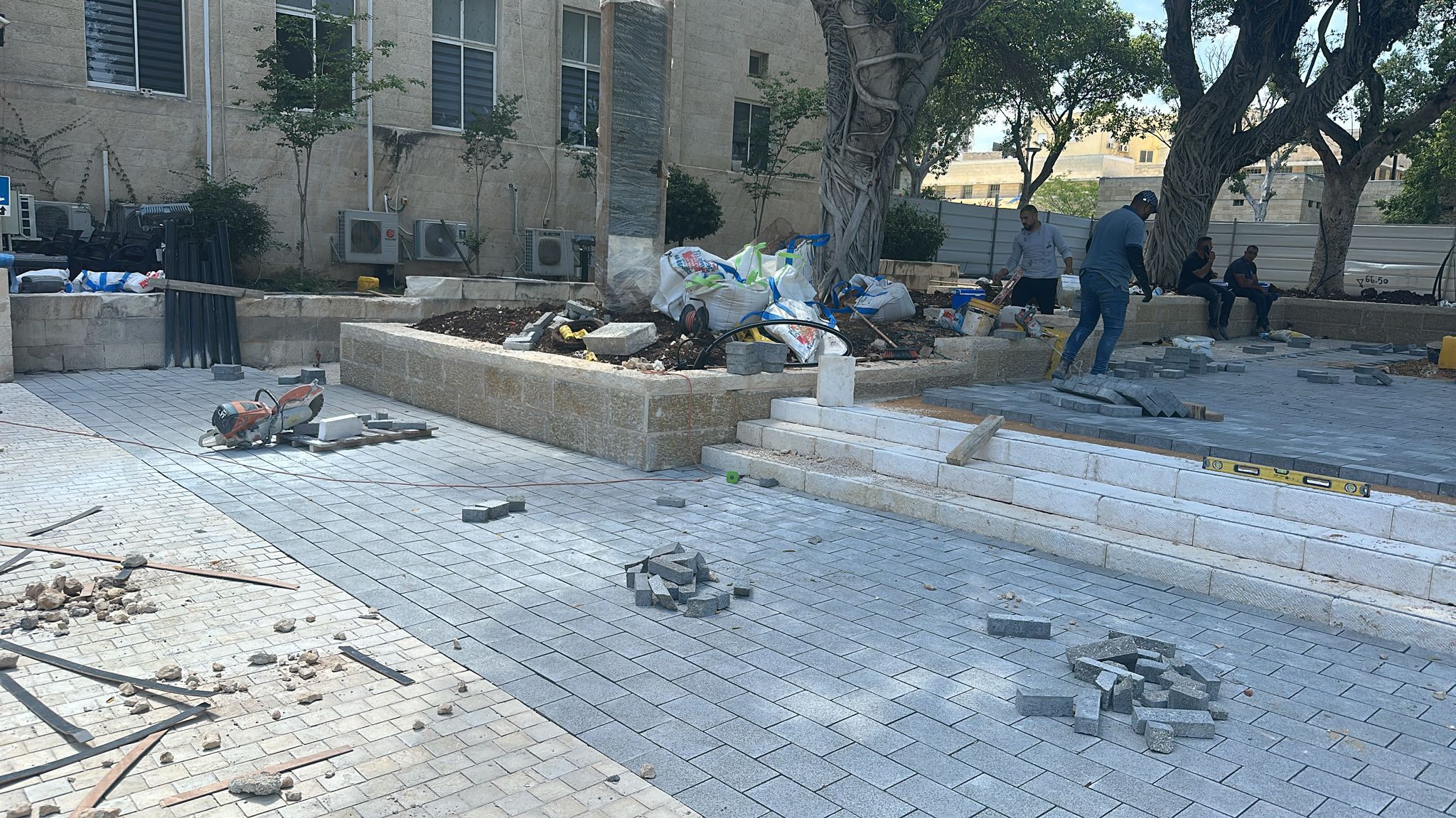 אתר בנייה שבו נראים פועלים המניחים אבני ריצוף אפורות, חומרים מפוזרים, כלים ופסולת, בסמוך לבניין אבן ועצים.