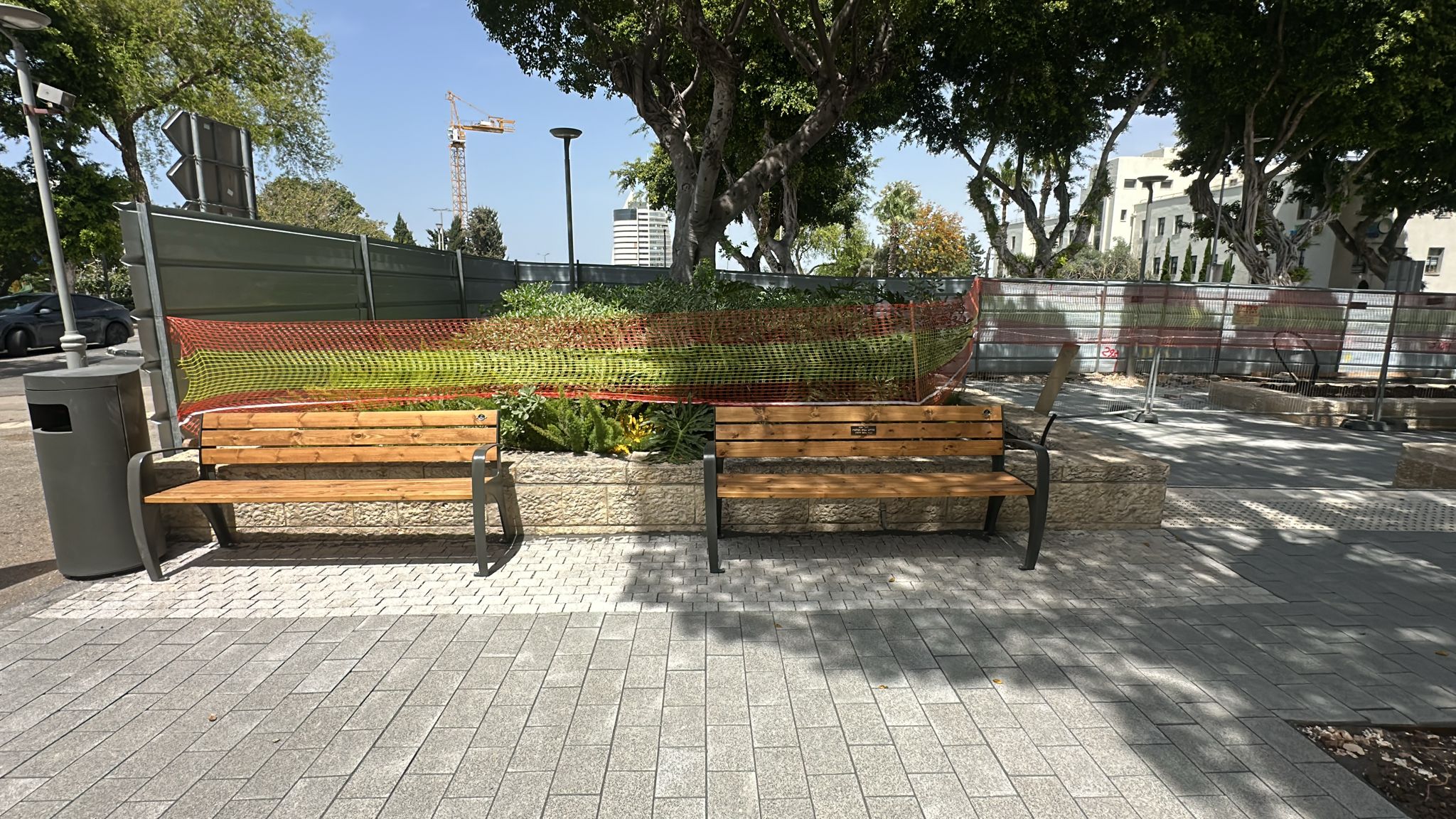 שני ספסלי עץ מוצבים על מדרכה מרוצפת ליד ערוגה עם צמחים, המוקפת ברשת בנייה כתומה ובגדר. ברקע נראים עצים ובניינים.