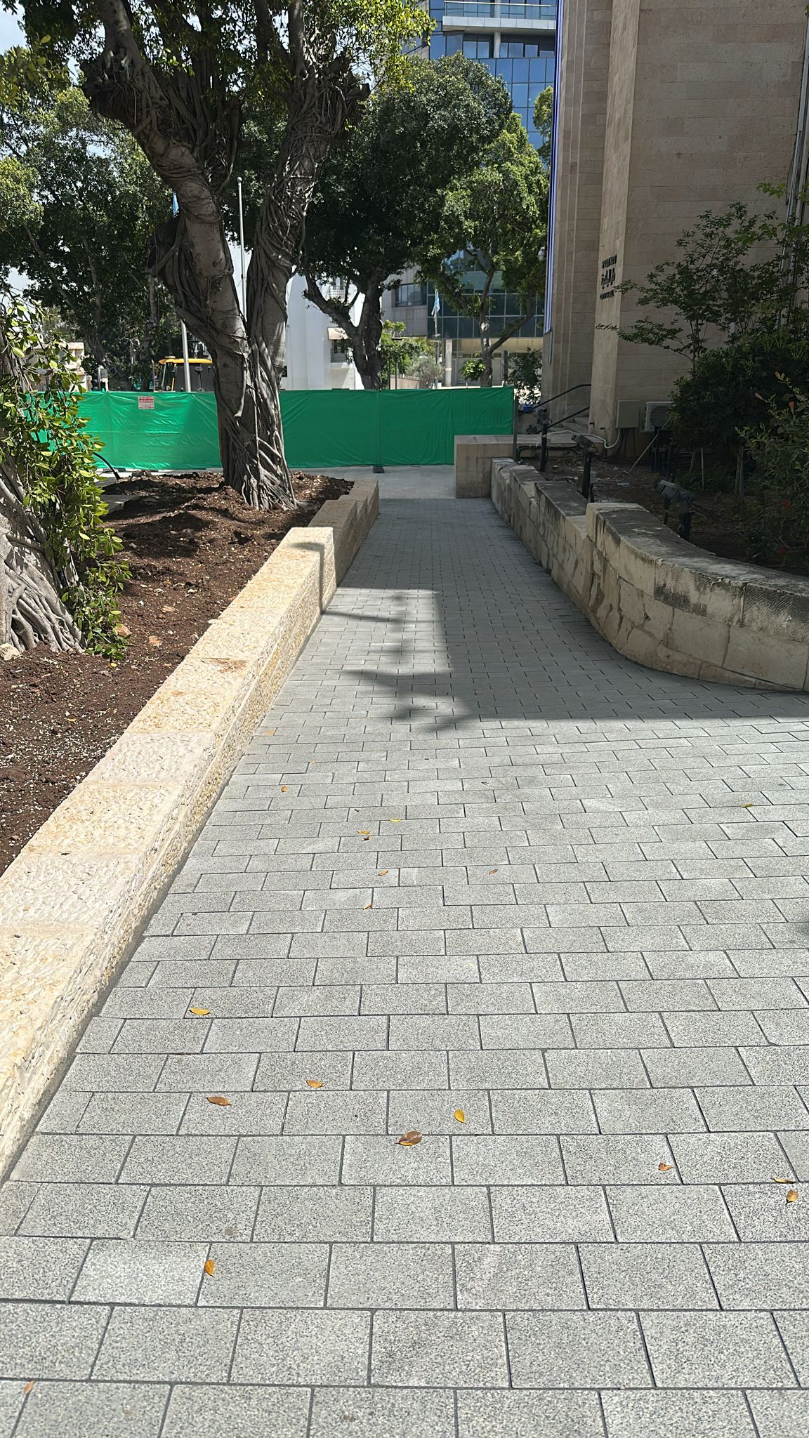 שביל מרוצף, המוקף בקירות אבן ועצים, מוביל לעבר גדר בנייה ירוקה ולבניין מודרני הנראה ברקע.