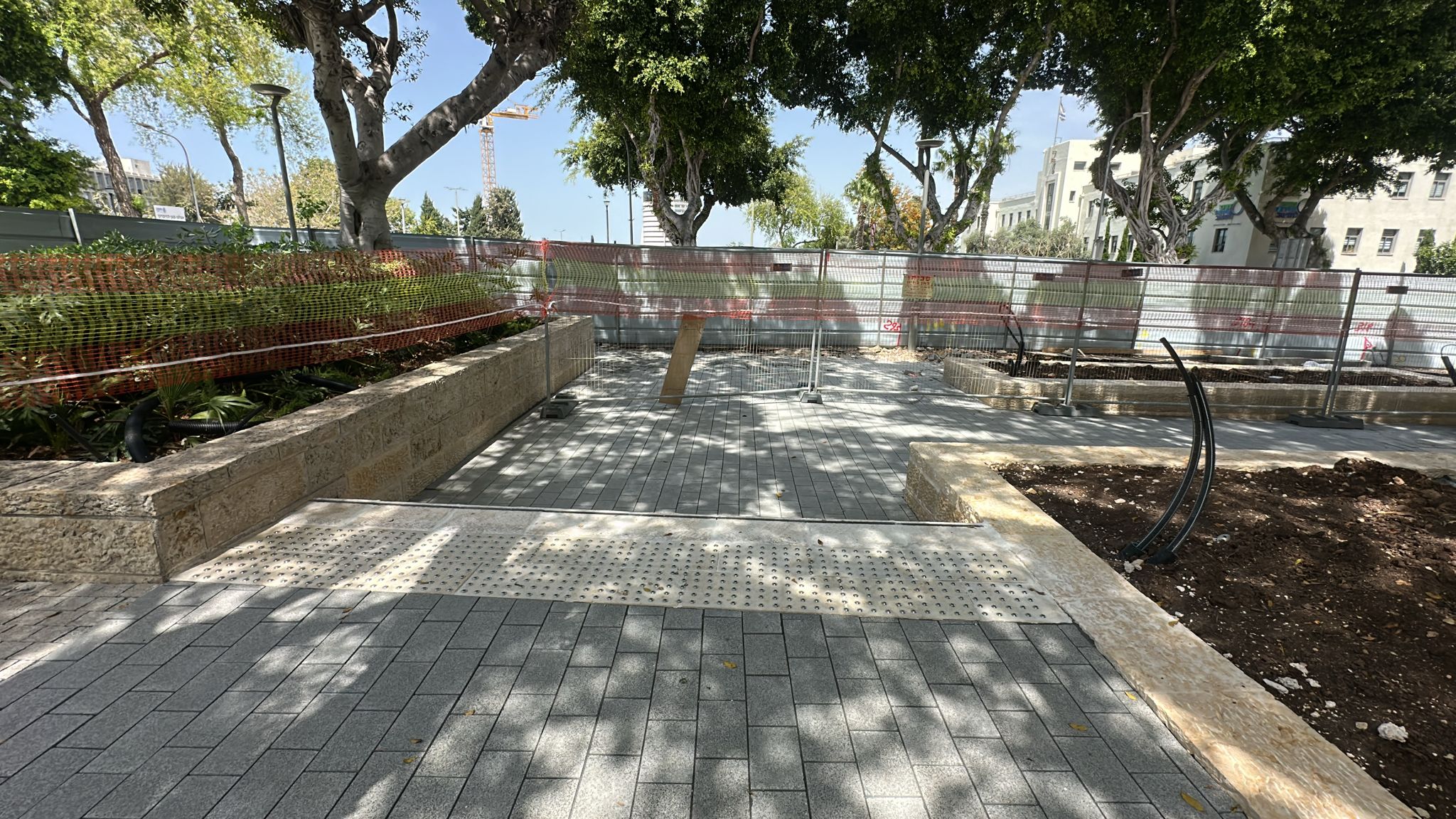 מדרכה מרוצפת עם רמפה וריצוף מובלט, המוקפת בעציצים וגדרות, תחת צל העצים ביום שטוף שמש.