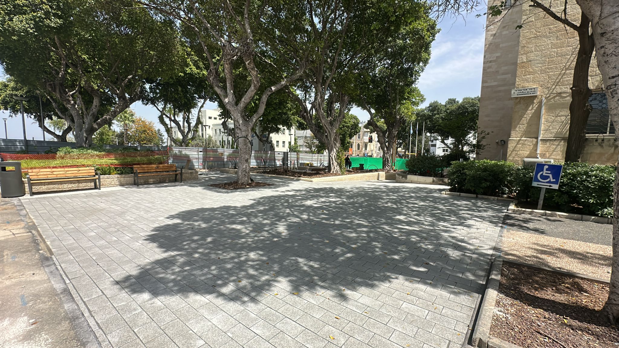 חצר מרוצפת עם ספסלים, עצים המספקים צל, מבנה מימין ושלט כחול המציין חניה לנכים.