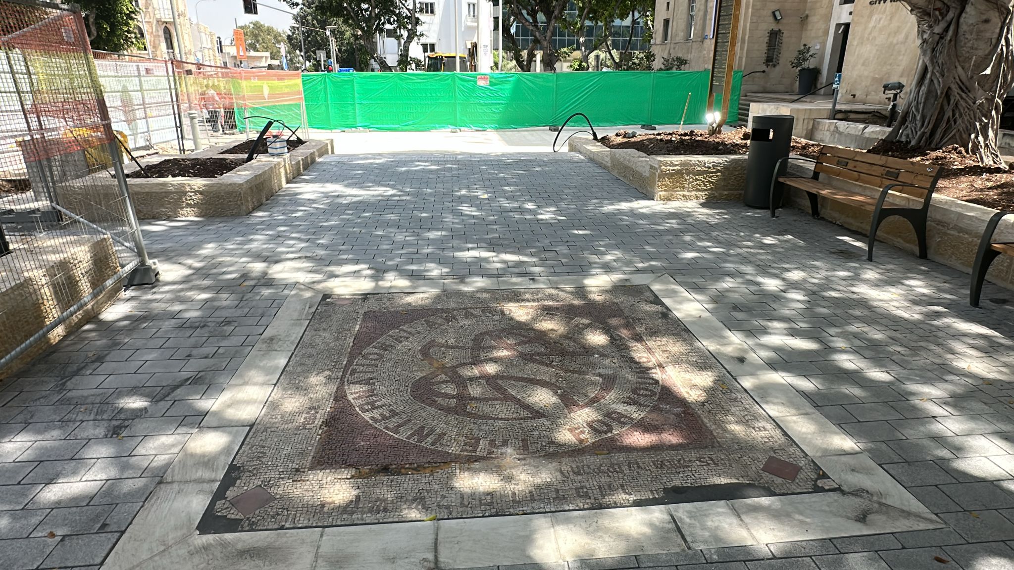שביל אבן רחב עם פסיפס גדול במרכזו, ספסלים ועצים מימין, וגדר בנייה ירוקה ברקע.