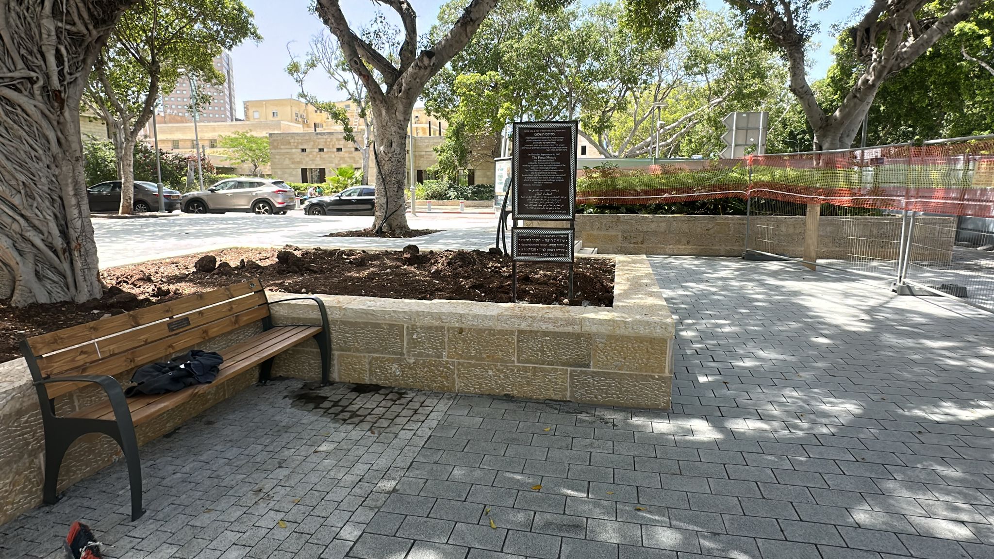 ספסל ליד פיסת אדמה ושלט על שטח מרוצף המוצל על ידי עצים, עם גדר רשת אדומה ומכוניות חונות ברקע.