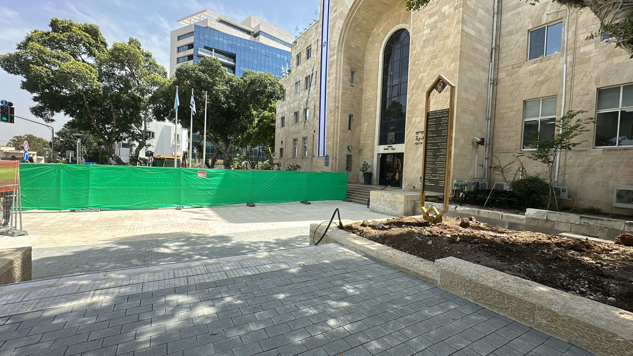גדר ירוקה חוסמת את הגישה לקטע המדרכה שמחוץ לבניין אבן בעל כניסה מקושתת; עצים מטילים צללים על השביל.