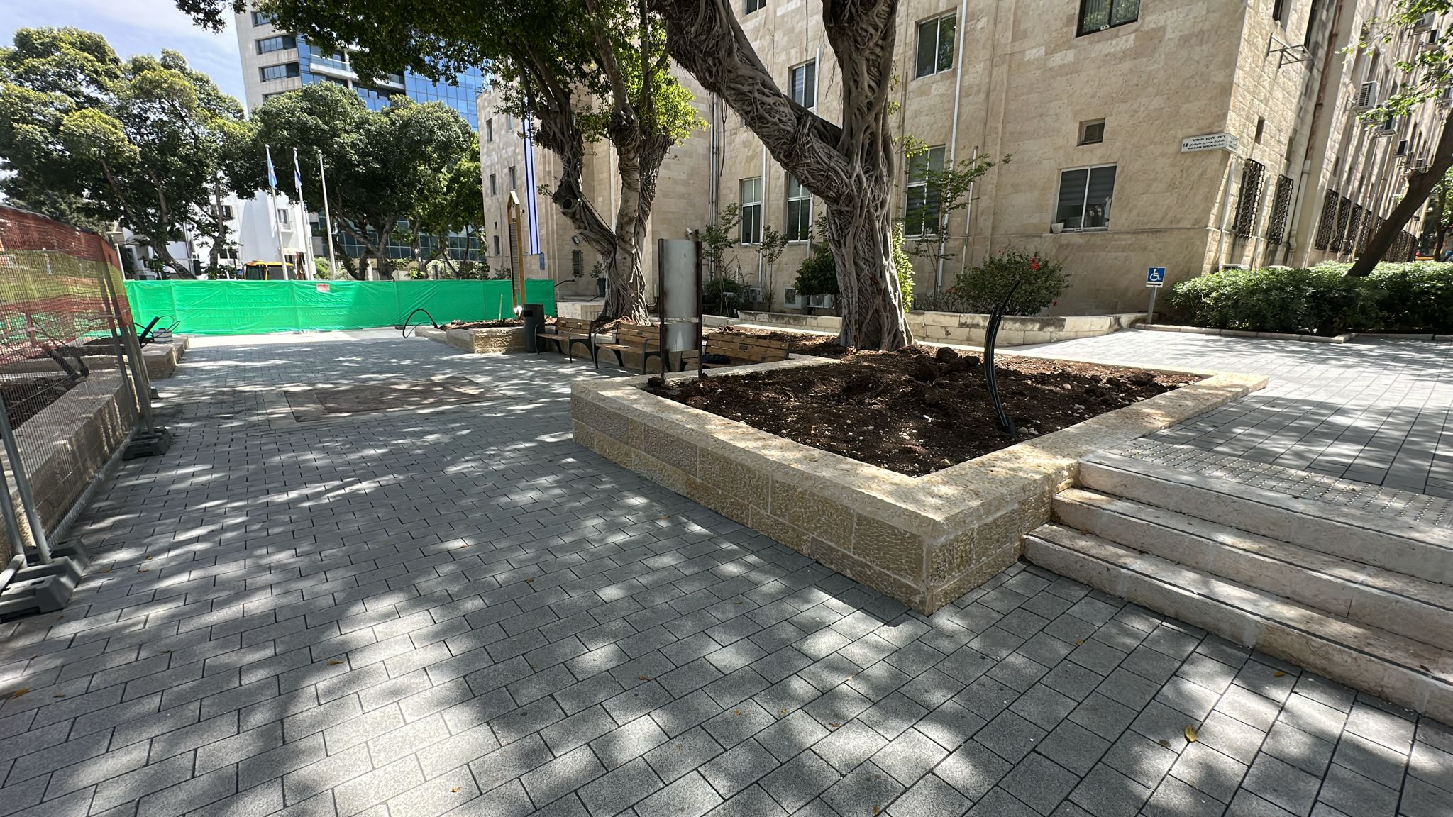 חצר עירונית מוארת בשמש, עם עציצים מאבן, עצים גדולים, ספסלים ומדרגות המובילות אל בניין. צללי העצים מכסים את המדרכה המעוטרת בדוגמאות. ברקע, גדר בנייה ירוקה.
